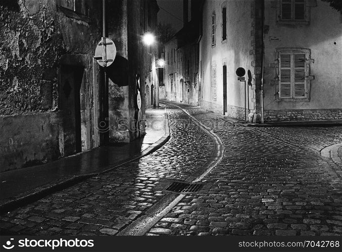 A night scene captures an empty street in the city of Beaune in France where the wet cobblestones are bright with reflected light from the street lights. (Scanned from black and white film.)