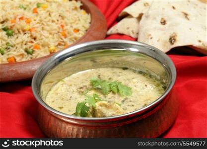 A old beaten copper serving bowl with homemade fish in green curry sauce, vegetable pilau rice on a terracotta plate and a pile of chappati indian flat breads.