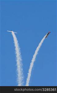 A pair of aerobatic airplanes trailing smoke flies separate in smooth arcs against a bright blue sky