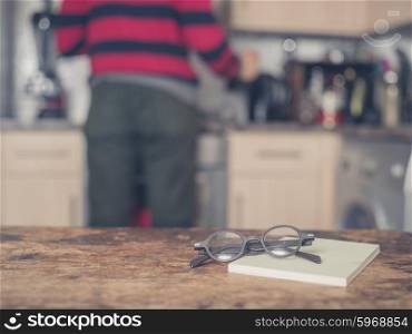 A pair of glasses and a book on a table in a kitchen. There is a man in the background