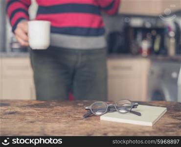 A pair of glasses and a book on a table in a kitchen. There is a man in the background with a cup of coffee