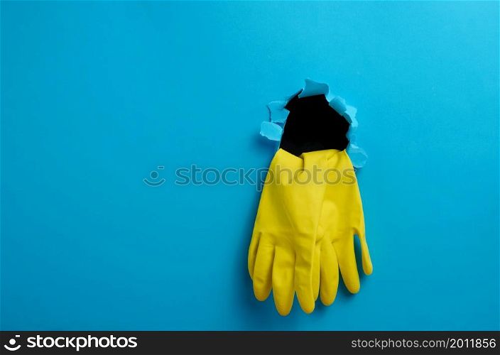 a pair of yellow latex house cleaning gloves sticking out of the torn hole of the blue paper background, copy space