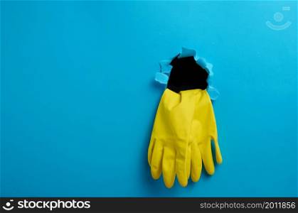a pair of yellow latex house cleaning gloves sticking out of the torn hole of the blue paper background, copy space