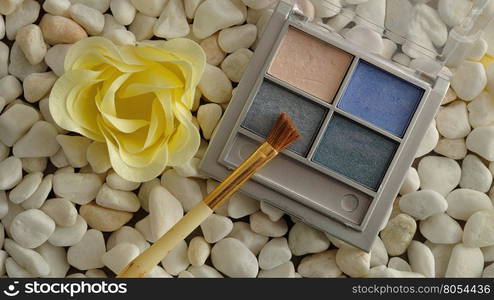 A palette with blue shades of eye shadow and a brush displayed on white pebbles with a white flower