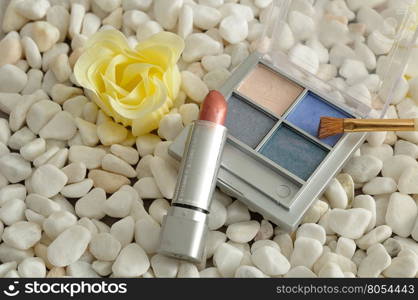 A palette with blue shades of eye shadow and a brush, with red lip stick displayed on white pebbles with a white flower