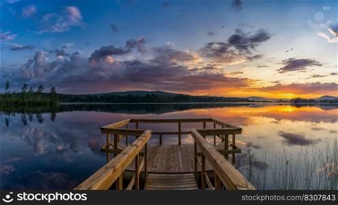 A panorama landscape of a wooden pier leads out onto a calm lake with forest on the opposite shore and a colorful sunset sky