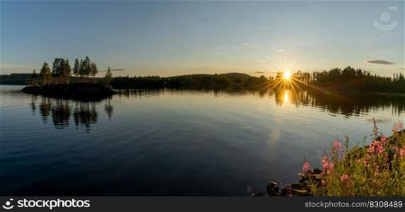 A panorama of a calm lake with small island and golden sunset evening light on the trees and forest on the lakeshore in the foreground and a sun star
