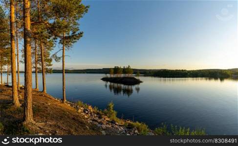 A panorama of a calm lake with small island and golden sunset evening light on the trees and forest on the lakeshore in the foreground