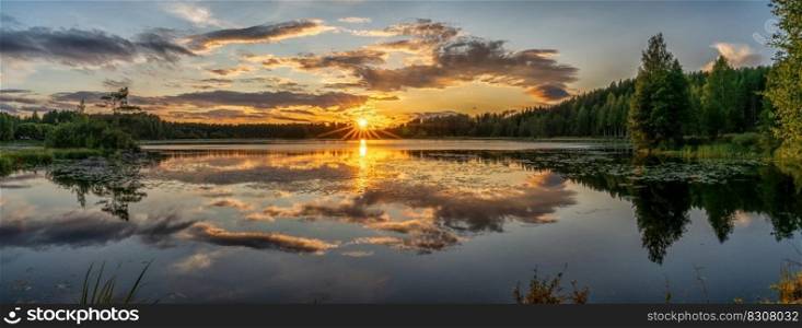A panorama of a colorful sunset reflected in a calm lake landscape with green forest and reeds