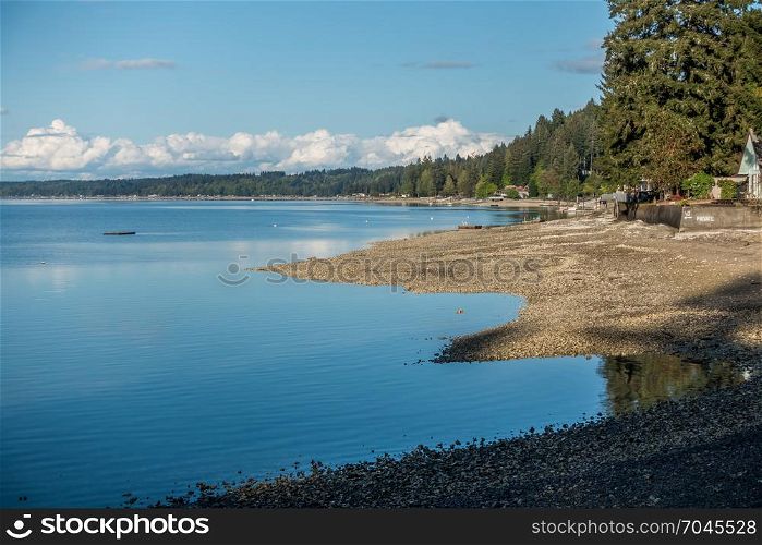 A panoramic veiw of homes along the coastline of Hood Canal in Washington State. The tide is low and the blue cloudy skies are reflected in the calm waters.