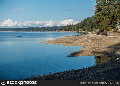 A panoramic veiw of homes along the coastline of Hood Canal in Washington State. The tide is low and the blue cloudy skies are reflected in the calm waters.