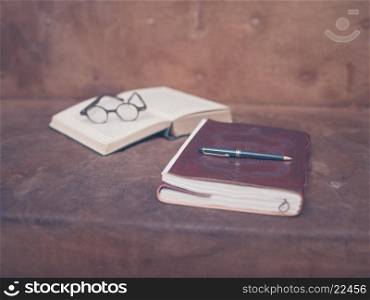 A pen on top of a notebook with an open book and a pair of glasses in the background