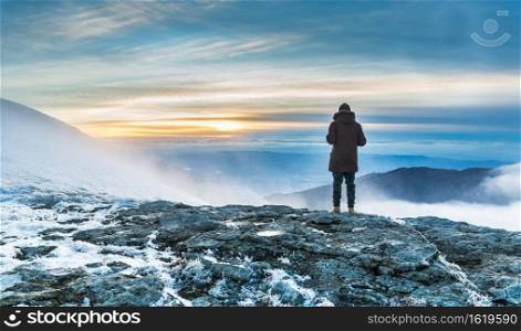 A person standing on a snow covered cliff over the breathtaking view of the mountains under the sunset. Person standing on a snow covered cliff over the breathtaking view of mountains under the sunset