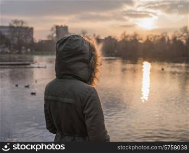 A person wearing a thick warm coat is standing by a pond in a park and is watching the sunset in winter
