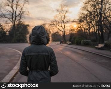 A person wearing a warm coat with a big hood is standing in a park at sunset