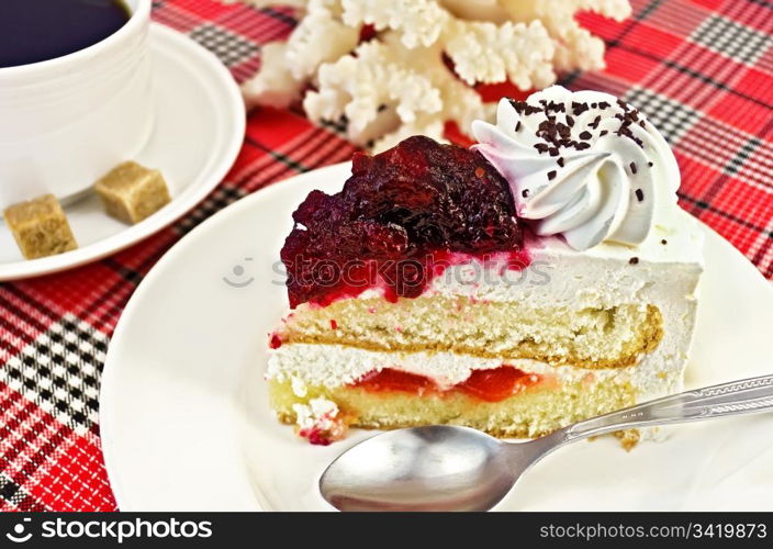 A piece of cake with white cream and red jelly on the plate, a white porcelain cup of coffee with two lumps of sugar, white coral on a background of red plaid fabric