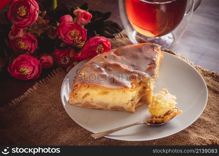 A piece of delicious apple strudel and a spoon on a plate. Amber tea in a transparent cup on a brown background. A piece of apple pie on a white plate on a background of pink flowers