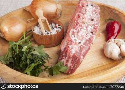 A piece of fresh marbled beef, chili pepper, parsley, onion, garlic, ribs lie on a wooden tray.