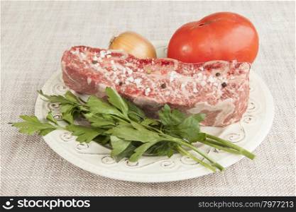 A piece of fresh marbled beef with sea salt and black pepper, tomato, onions on a porcelain plate.