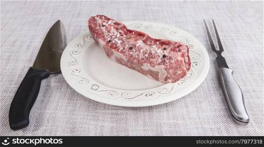 A piece of fresh marbled beef with sea salt and black pepper, knife and fork on a porcelain plate.