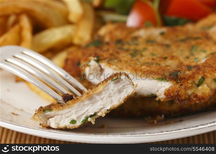 A piece of homemade breaded chicken schnitzel or escalope on a fork with french fries and a tomato and green salad behind