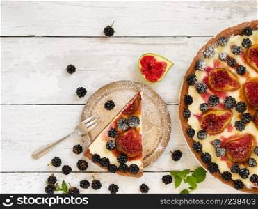 A piece of homemade pie with figs and blackberries is located on a vintage plate. Nearby is a pie. Wooden vintage background. Next to the pie are blackberries, cut figs and cutlery. Top view.
