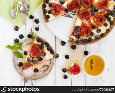 A piece of homemade pie with figs and blackberries is located on a vintage plate. Nearby is a pie and a cup of green tea. Wooden vintage background. Next to the pie are blackberries, cut figs and cutlery. Top view.