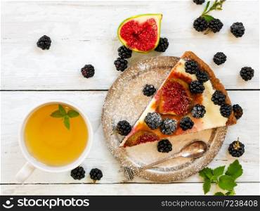 A piece of homemade pie with figs and blackberries is located on a vintage plate. Nearby is a cup of green tea. Wooden vintage background. Next to the pie are blackberries, cut figs and cutlery. Top view.. A piece of homemade pie with figs and blackberries is located on a vintage plate. Nearby is a cup of green tea with mint. Wooden vintage background. Next to the pie are blackberries, cut figs and cutlery. Top view.