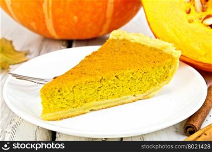 A piece of pumpkin pie with whipped cream, a fork in the white plate, pumpkin and cinnamon on a background of bright wooden planks