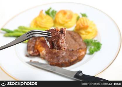 A piece of ribeye steak on a fork with the meal of steak, asparagus and duchesse potatoes out of focus in the background.