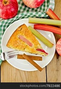 A piece of sweet cake with rhubarb and apples, cinnamon, napkin, fork on a wooden boards background