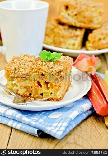 A piece of sweet cake with rhubarb, mint, blue napkin, cup on the background of wooden boards