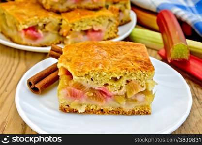 A piece of sweet cake with rhubarb, rhubarb stalks, cinnamon, blue napkin on background of wooden boards