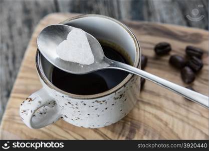 A piece of white sugar in the form of heart lies on a teaspoon over a cup of black coffee