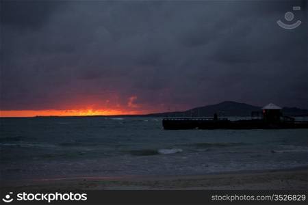 A pier against the horizon with a fiery birght red and orange sunset as the light goes down over the horizon in the Galapagos Islands of Ecaudor