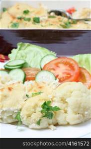 A plate of cauliflower cheese or gratin served with a salad of tomato, lettuce and cucumber, with the serving bowl in the background
