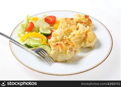 A plate of cauliflower cheese served with a fork and a salad of lettuce, cucumber, tomato and capsicum, a traditional English dish.
