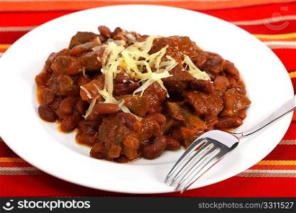 A plate of chili con carne made with red kidney beans and lumps of steak, topped with grated cheddar cheese, with a fork on a table