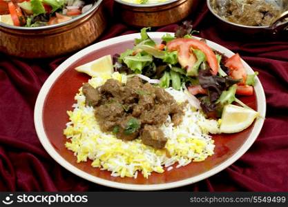 A plate of homemade beef rogan josh, served with yellow and white rice and a salad, with typical Indian serving bowls behind. Rogan josh is usually made with lamb, as it is a Hindu dish, but works equally well with beef.