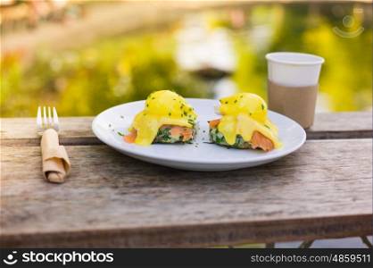 A plate with eggs benedict made with salmon and hollandaise sauce on a table by a pond in the park