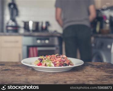 A plate with salad on a table in a kitchen with a man cooking in the background