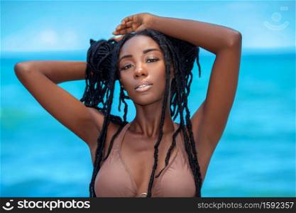 A portrait of a lascivious attractive young black female with beautiful makeup & long dreadlocks posing by herself on a sunny summer day at a tropical beach wearing a brown bikini top.