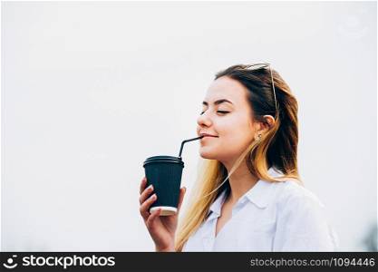 a portrait of a pretty long haired young girl wearing white shirt, drinking coffee or smoothie through a bendie, smiling, eyes closed, outdoors. Copy space