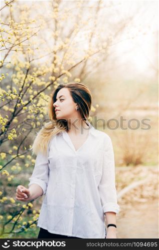 A portrait of a pretty long haired young woman in a garden enjoying blooming trees with yellow flowers, romantically looking to the side, film grain effect