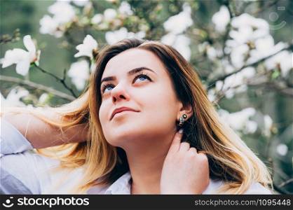 A portrait of a pretty young girl in a garden enjoying blooming magnolia trees with white flowers, romantically looking up, closeup view, blurred background