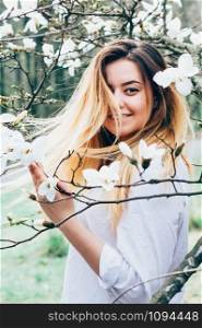 A portrait of a pretty young long haired girl in a garden enjoying blooming magnolia trees with white flowers, romantic look, closeup view, blurred background and foreground with branches, her hair in motion