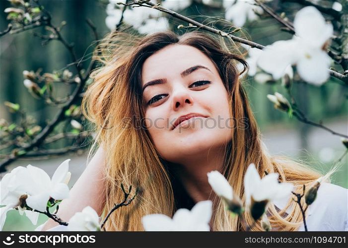 A portrait of a pretty young long haired girl in a garden enjoying blooming magnolia trees with white flowers, romantic look, closeup view, blurred background and foreground with branches