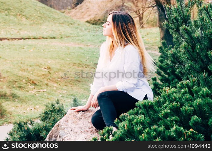 A portrait of a pretty young long haired girl in white shirt, sitting on a stone in a park, with green grass background and pine trees in the foreground