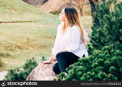 A portrait of a pretty young long haired girl in white shirt, sitting on a stone in a park, with green grass background and pine trees in the foreground