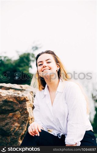 A portrait of a pretty young long haired girl in white shirt, smiling, sitting on a big stone in a park, with green trees in the background